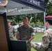 Military personnel from multiple branches are gathered under a black canopy tent at an outdoor event. A U.S. Marine in MARPAT camouflage is being briefed or presented to by a soldier wearing a maroon beret, while another soldier in a green beret stands in the background. The tent banner reads 'Security Force Assistance' and 'Forward!'