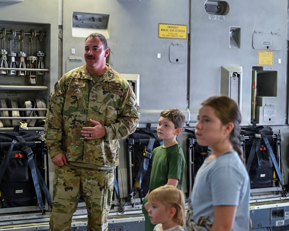 A U.S. Air Force service member in OCP (Operational Camouflage Pattern) uniform, identified by a name tape reading 'MURPHY,' is speaking to a group of children inside what appears to be a military cargo aircraft, likely a C-17 Globemaster III. The setting suggests a familiarization tour or 'Families Day' event where children are being shown the interior of the aircraft. The service member appears to be explaining aircraft systems or equipment to the attentive children.