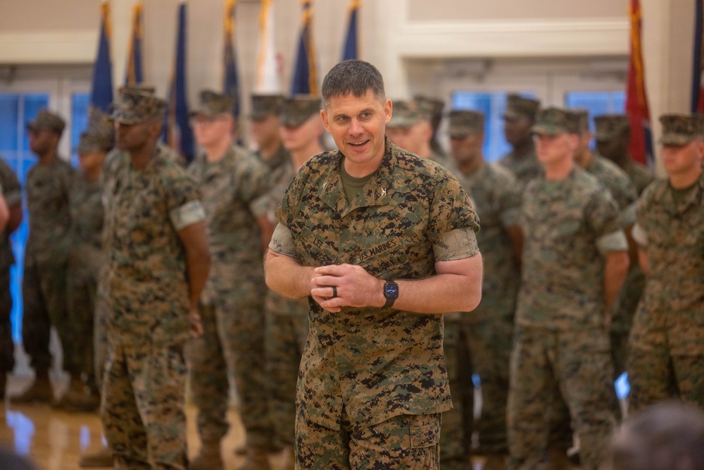 A U.S. Marines officer stands in the foreground of an indoor military ceremony or formation, smiling with hands clasped together. He is wearing MARPAT woodland digital camouflage uniform with 'U.S. MARINES' tape visible on his chest. Behind him, numerous Marines in similar uniforms stand in formation, with multiple military flags/guidons visible in the background.