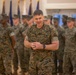 A U.S. Marines officer stands in the foreground of an indoor military ceremony or formation, smiling with hands clasped together. He is wearing MARPAT woodland digital camouflage uniform with 'U.S. MARINES' tape visible on his chest. Behind him, numerous Marines in similar uniforms stand in formation, with multiple military flags/guidons visible in the background.