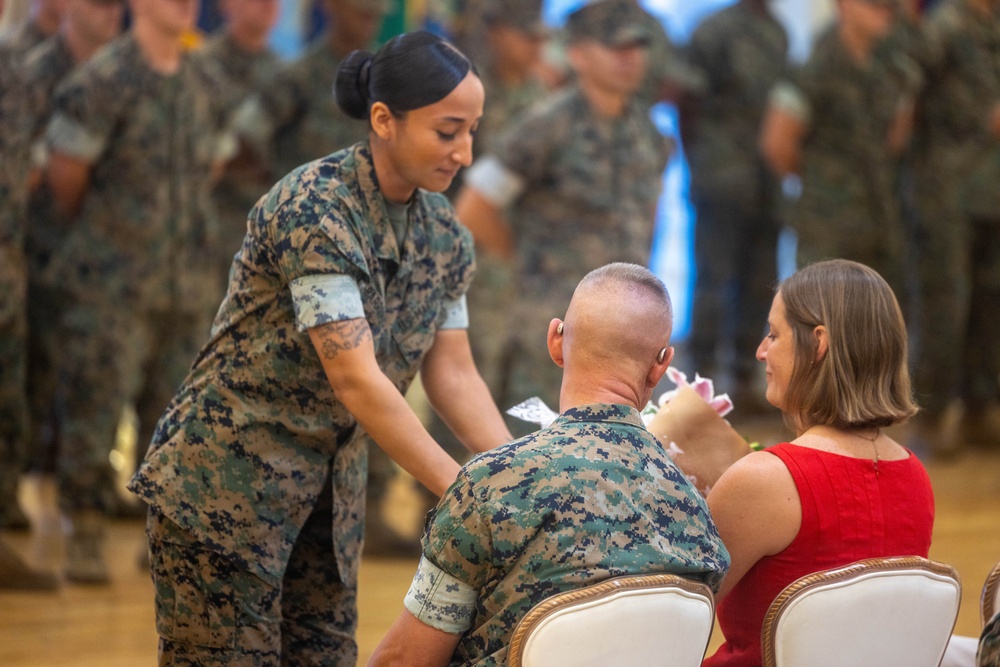 A military ceremony is taking place in an indoor venue, likely a formal hall. A female service member in MARPAT camouflage uniform is leaning forward to present or pin something to a seated male service member, while a civilian woman in a red dress sits beside him. Rows of uniformed personnel stand in formation in the background.