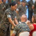 A military ceremony is taking place in an indoor venue, likely a formal hall. A female service member in MARPAT camouflage uniform is leaning forward to present or pin something to a seated male service member, while a civilian woman in a red dress sits beside him. Rows of uniformed personnel stand in formation in the background.