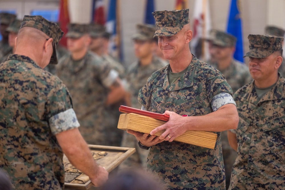 A formal U.S. Marines military ceremony is taking place indoors, likely a retirement or award ceremony. A central Marine in MARPAT camouflage uniform is holding a wooden presentation box containing what appears to be red-covered documents or certificates. Multiple Marines in uniform are present in the background alongside several military branch flags.