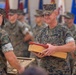 A formal U.S. Marines military ceremony is taking place indoors, likely a retirement or award ceremony. A central Marine in MARPAT camouflage uniform is holding a wooden presentation box containing what appears to be red-covered documents or certificates. Multiple Marines in uniform are present in the background alongside several military branch flags.