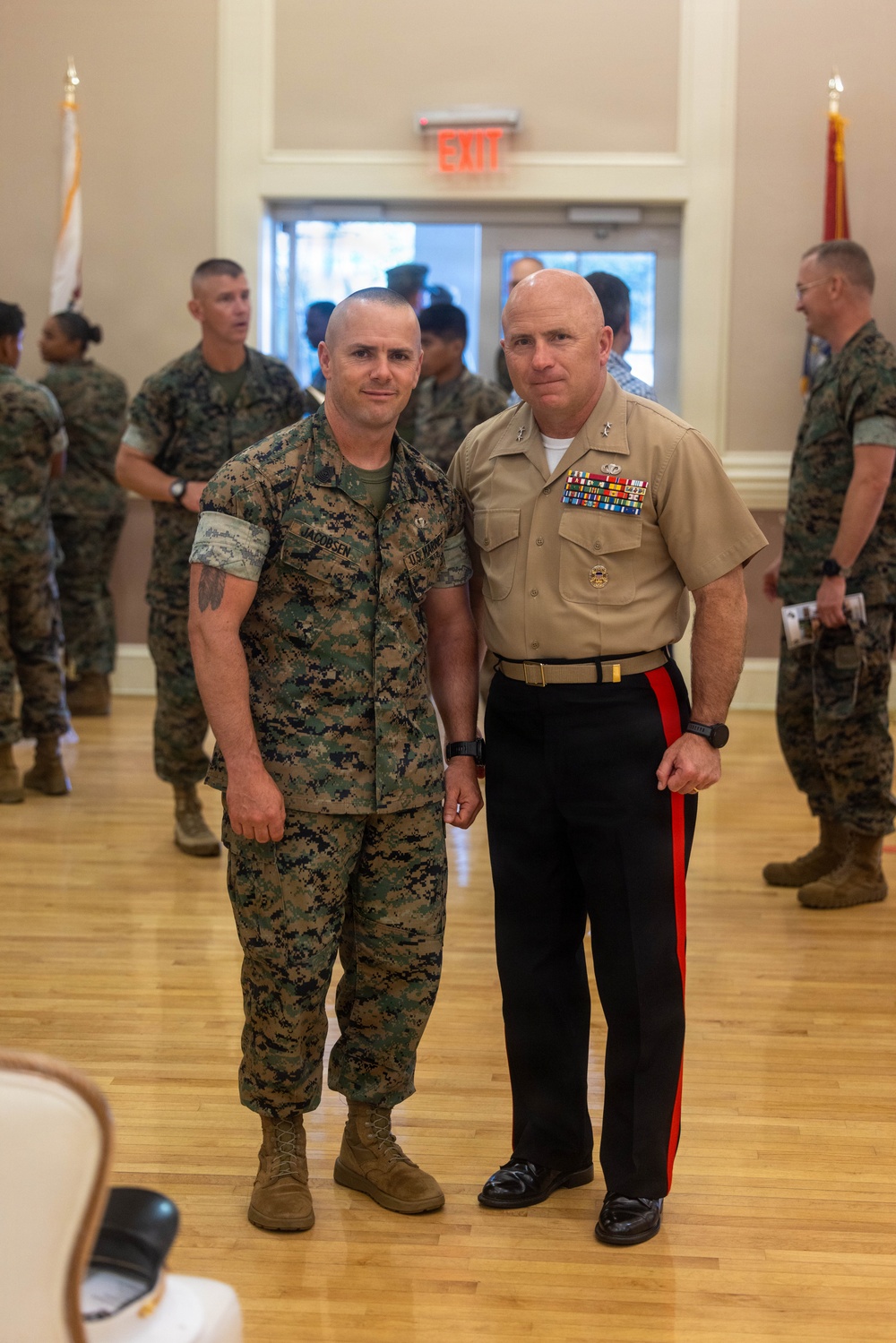 Two U.S. Marines pose together in an indoor ceremonial setting, likely during a formal military event or ceremony. The marine on the left wears MARPAT camouflage utilities with a name tape reading 'JACOBSEN,' while the senior officer on the right wears the tan Service 'C' uniform with black trousers featuring a red blood stripe, indicating a senior enlisted or officer rank. Multiple other Marines in camouflage utilities are visible in the background.