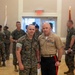 Two U.S. Marines pose together in an indoor ceremonial setting, likely during a formal military event or ceremony. The marine on the left wears MARPAT camouflage utilities with a name tape reading 'JACOBSEN,' while the senior officer on the right wears the tan Service 'C' uniform with black trousers featuring a red blood stripe, indicating a senior enlisted or officer rank. Multiple other Marines in camouflage utilities are visible in the background.