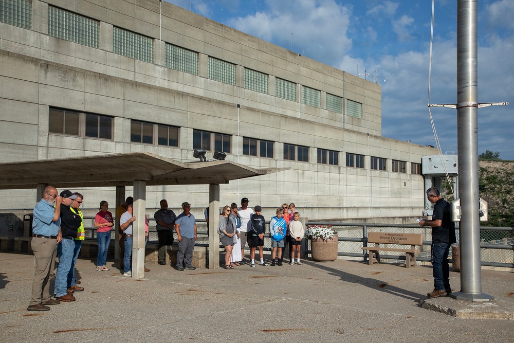 A small outdoor memorial ceremony is taking place at what appears to be a dam or industrial facility. A group of approximately 15 people are gathered near a bench and flower planter dedicated 'In Memoriam of Bruce Dvoracek,' while a man on the right appears to be reading from a document. The setting is a concrete plaza in front of a large brutalist-style building on a sunny day.