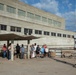 A small outdoor memorial ceremony is taking place at what appears to be a dam or industrial facility. A group of approximately 15 people are gathered near a bench and flower planter dedicated 'In Memoriam of Bruce Dvoracek,' while a man on the right appears to be reading from a document. The setting is a concrete plaza in front of a large brutalist-style building on a sunny day.