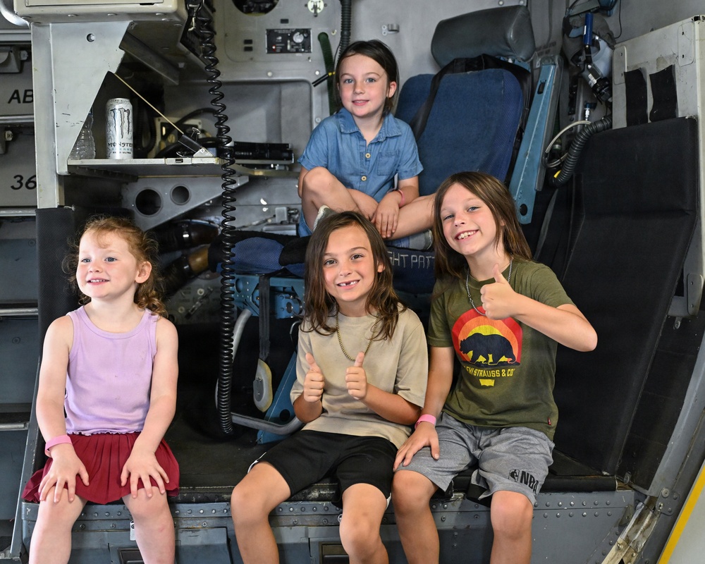 Four children are posing inside what appears to be a military aircraft or helicopter, likely during a public airshow or museum visit. Three children sit on a lower ledge while one sits elevated on a seat behind them. Two of the children are giving thumbs-up gestures, suggesting excitement and enjoyment.