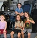 Four children are posing inside what appears to be a military aircraft or helicopter, likely during a public airshow or museum visit. Three children sit on a lower ledge while one sits elevated on a seat behind them. Two of the children are giving thumbs-up gestures, suggesting excitement and enjoyment.