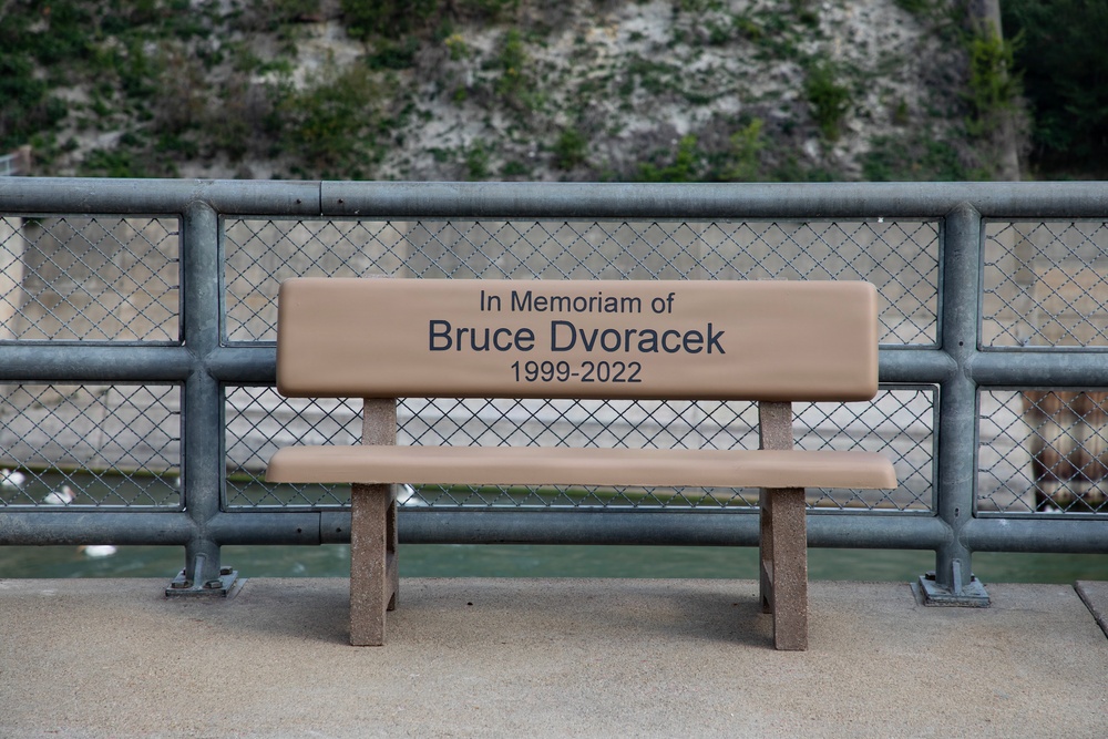 A memorial bench dedicated to Bruce Dvoracek (1999–2022) is positioned along a waterfront walkway or bridge. The bench features an engraved dedication on its backrest reading 'In Memoriam of Bruce Dvoracek 1999-2022'. The setting includes a chain-link and metal railing fence with a rocky, vegetated cliff visible in the background.