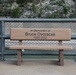 A memorial bench dedicated to Bruce Dvoracek (1999–2022) is positioned along a waterfront walkway or bridge. The bench features an engraved dedication on its backrest reading 'In Memoriam of Bruce Dvoracek 1999-2022'. The setting includes a chain-link and metal railing fence with a rocky, vegetated cliff visible in the background.