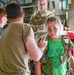A young girl wearing a green t-shirt is being fitted with a tactical harness/parachute rig by a person in a tan military-style shirt inside what appears to be a military aircraft. A soldier in camouflage uniform observes in the background, along with another child in a black shirt. This appears to be an educational or familiarization event where children are being shown military equipment.