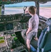 A young boy stands in the cockpit of a large military aircraft, looking out the front windshield at the airfield. The cockpit is filled with active instrumentation including digital displays, navigation screens, and numerous switches and controls. A placard on the pilot's seat references a life vest, and tail markings suggest this is a U.S. Air Force C-17 Globemaster III.