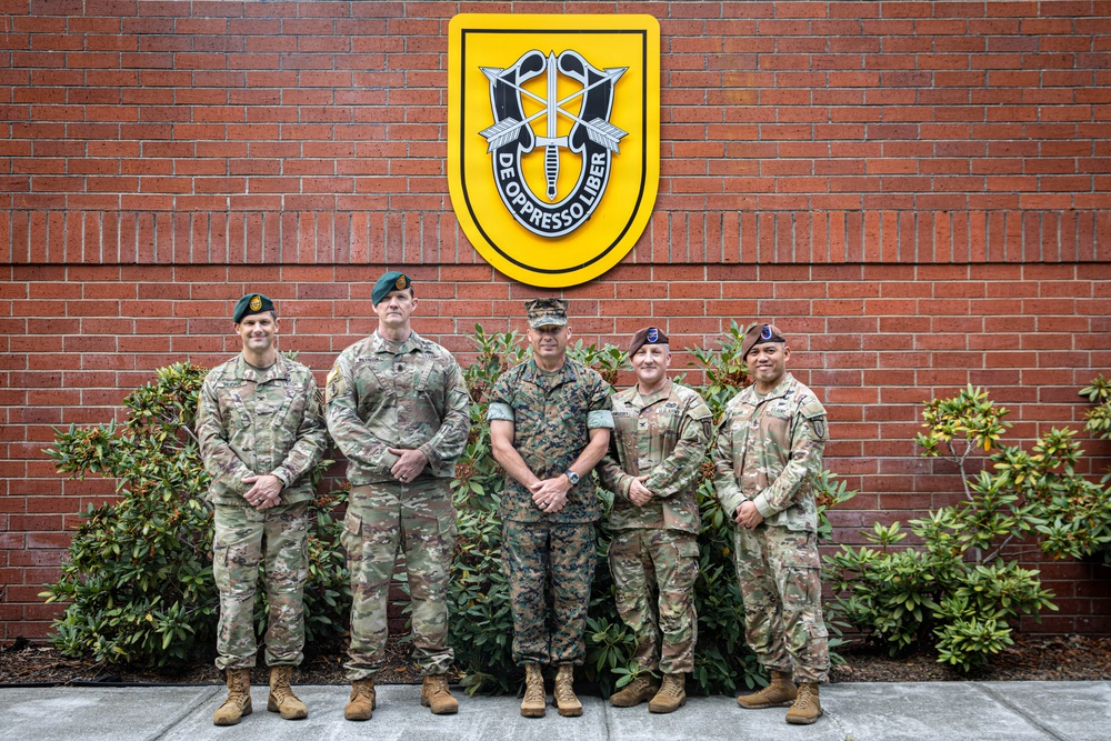 Five military personnel pose for a formal group photo in front of a brick wall displaying the U.S. Army Special Forces insignia with the motto 'De Oppresso Liber.' The group includes members from different branches, identifiable by their distinct camouflage uniforms. The setting appears to be an official military installation.