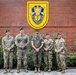 Five military personnel pose for a formal group photo in front of a brick wall displaying the U.S. Army Special Forces insignia with the motto 'De Oppresso Liber.' The group includes members from different branches, identifiable by their distinct camouflage uniforms. The setting appears to be an official military installation.