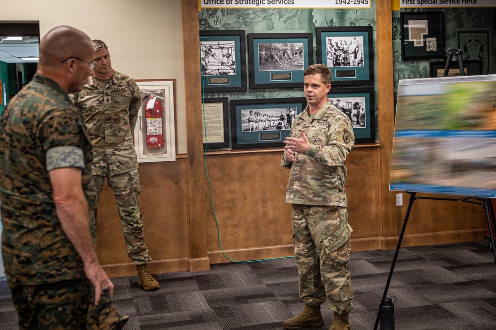 Three military personnel are gathered in what appears to be a museum or briefing room featuring historical displays about the Office of Strategic Services (1942-1945) and the First Special Service Force. One soldier in Army multicam uniform is actively speaking and gesturing, while the other two — one in Marine Corps MARPAT and one in Army multicam — are listening. A presentation board with images is visible to the right.