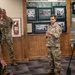 Three military personnel are gathered in what appears to be a museum or briefing room featuring historical displays about the Office of Strategic Services (1942-1945) and the First Special Service Force. One soldier in Army multicam uniform is actively speaking and gesturing, while the other two — one in Marine Corps MARPAT and one in Army multicam — are listening. A presentation board with images is visible to the right.