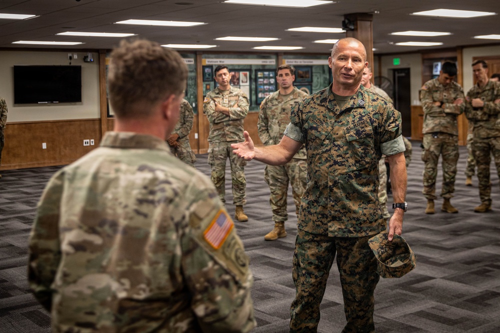 A U.S. Marine officer in MARPAT digital camouflage is engaged in conversation with a U.S. Army soldier in OCP (Operational Camouflage Pattern) uniform inside what appears to be a military briefing or conference room. The Marine is gesturing with one hand while holding his cover (cap) in the other, suggesting an informal discussion or briefing. Several other Army soldiers in OCP uniforms observe the interaction in the background.