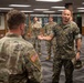 A U.S. Marine officer in MARPAT digital camouflage is engaged in conversation with a U.S. Army soldier in OCP (Operational Camouflage Pattern) uniform inside what appears to be a military briefing or conference room. The Marine is gesturing with one hand while holding his cover (cap) in the other, suggesting an informal discussion or briefing. Several other Army soldiers in OCP uniforms observe the interaction in the background.