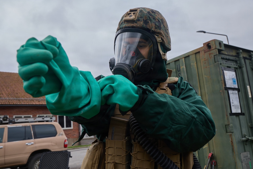 A military service member is donning CBRN (Chemical, Biological, Radiological, and Nuclear) protective gear, including a full-face gas mask, tactical helmet with digital camouflage cover, and bright green chemical-resistant gloves. The individual appears to be pulling on or adjusting the gloves as part of a suit-up procedure. The setting appears to be a military installation or base, with a green military container and a tan vehicle visible in the background.