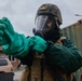 A military service member is donning CBRN (Chemical, Biological, Radiological, and Nuclear) protective gear, including a full-face gas mask, tactical helmet with digital camouflage cover, and bright green chemical-resistant gloves. The individual appears to be pulling on or adjusting the gloves as part of a suit-up procedure. The setting appears to be a military installation or base, with a green military container and a tan vehicle visible in the background.
