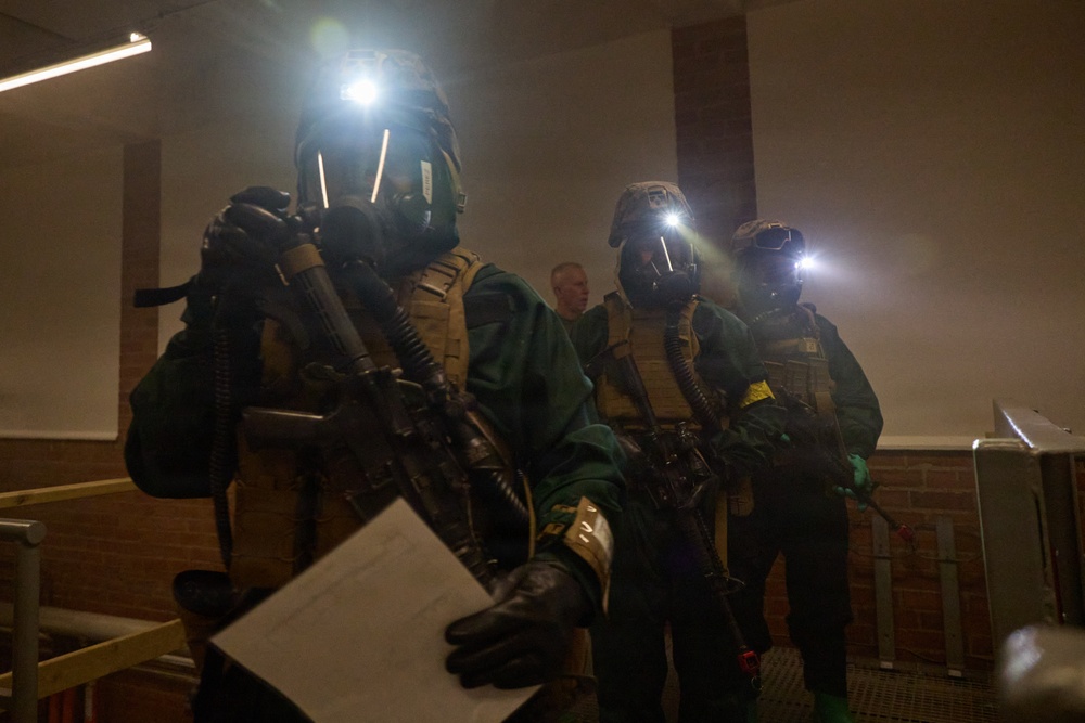Three heavily equipped personnel wearing gas masks, tactical vests, and carrying rifles are positioned inside what appears to be a building interior. Each individual has an active helmet-mounted light source illuminating the dimly lit environment. One person in the foreground is holding a document or paper while gripping a firearm.