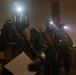 Three heavily equipped personnel wearing gas masks, tactical vests, and carrying rifles are positioned inside what appears to be a building interior. Each individual has an active helmet-mounted light source illuminating the dimly lit environment. One person in the foreground is holding a document or paper while gripping a firearm.