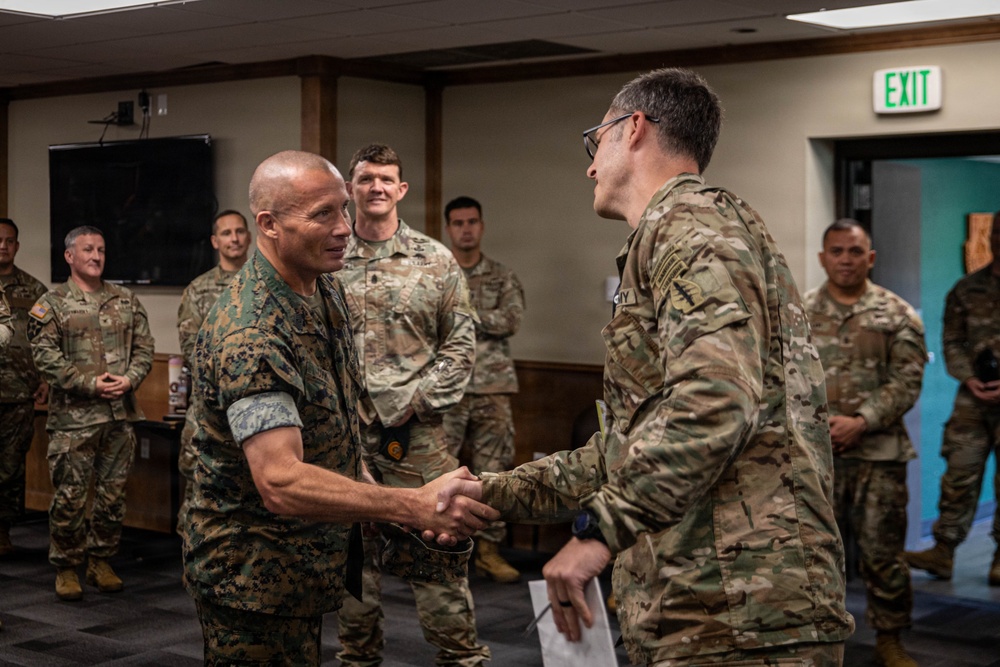 A formal military ceremony or meeting is taking place indoors, where a Marine Corps officer in MARPAT digital camouflage is shaking hands with an Army soldier in OCP (Operational Camouflage Pattern) uniform. Multiple military personnel from what appears to be both the U.S. Marine Corps and U.S. Army are gathered in the background observing the interaction. The setting appears to be a conference or briefing room on a military installation.