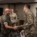 A formal military ceremony or meeting is taking place indoors, where a Marine Corps officer in MARPAT digital camouflage is shaking hands with an Army soldier in OCP (Operational Camouflage Pattern) uniform. Multiple military personnel from what appears to be both the U.S. Marine Corps and U.S. Army are gathered in the background observing the interaction. The setting appears to be a conference or briefing room on a military installation.