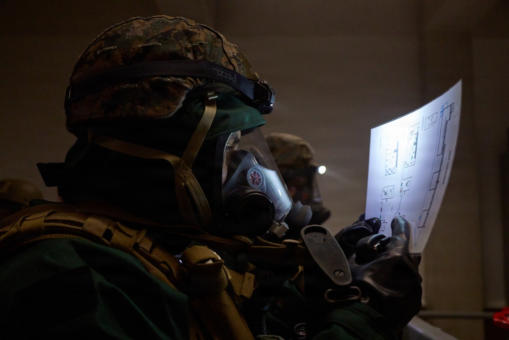 A military personnel wearing full CBRN (Chemical, Biological, Radiological, Nuclear) protective gear, including a gas mask and combat helmet with a Petzl headlamp, reviews what appears to be a building floor plan or schematic document. The environment is dark and smoky, suggesting a training exercise or operational scenario. A second similarly equipped individual is visible in the background.