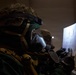 A military personnel wearing full CBRN (Chemical, Biological, Radiological, Nuclear) protective gear, including a gas mask and combat helmet with a Petzl headlamp, reviews what appears to be a building floor plan or schematic document. The environment is dark and smoky, suggesting a training exercise or operational scenario. A second similarly equipped individual is visible in the background.