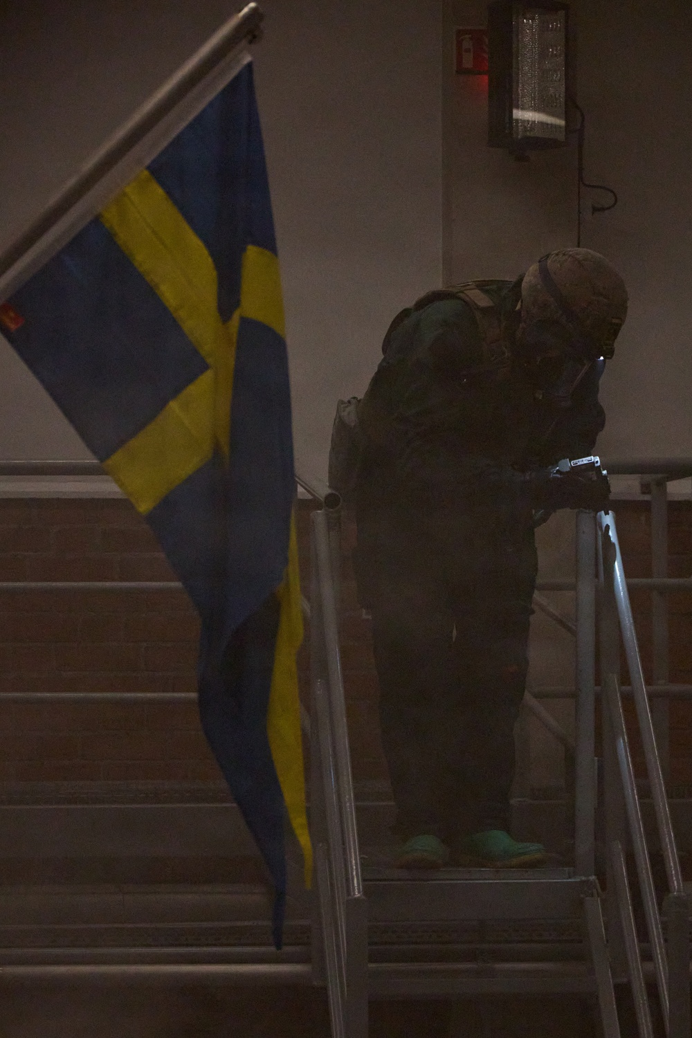 A heavily equipped individual in tactical/military gear is positioned on a staircase next to a Swedish flag in a dimly lit interior environment. The person appears to be in a crouched or leaning posture, holding what appears to be a flashlight or weapon-mounted light. The scene suggests a military exercise, security drill, or tactical training scenario.