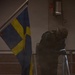 A heavily equipped individual in tactical/military gear is positioned on a staircase next to a Swedish flag in a dimly lit interior environment. The person appears to be in a crouched or leaning posture, holding what appears to be a flashlight or weapon-mounted light. The scene suggests a military exercise, security drill, or tactical training scenario.