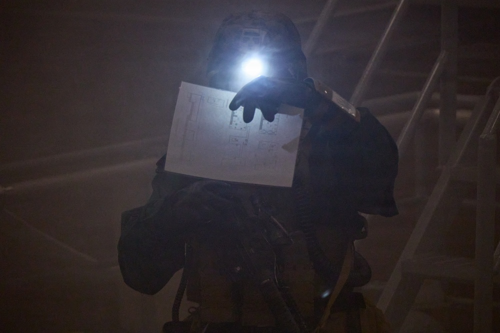A person wearing full protective gear, including a helmet with an active headlamp, is holding up a floor plan or architectural diagram in a dark, smoke-filled environment. The individual appears to be a first responder or tactical operator navigating a low-visibility situation. The headlamp illuminates the document being reviewed, suggesting active navigation or search operations.