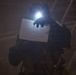 A person wearing full protective gear, including a helmet with an active headlamp, is holding up a floor plan or architectural diagram in a dark, smoke-filled environment. The individual appears to be a first responder or tactical operator navigating a low-visibility situation. The headlamp illuminates the document being reviewed, suggesting active navigation or search operations.