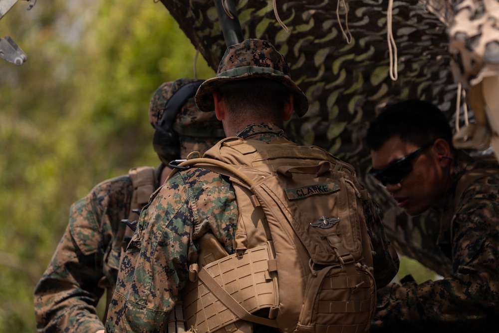 The image shows multiple military personnel in woodland MARPAT (Marine Pattern) camouflage uniforms working together near camouflage-netted equipment in an outdoor setting. The central figure, identified by a name tape as 'CLARKE,' is wearing a tan tactical backpack with a military badge visible and a boonie hat. The personnel appear to be engaged in a field operation or training exercise.