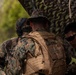 The image shows multiple military personnel in woodland MARPAT (Marine Pattern) camouflage uniforms working together near camouflage-netted equipment in an outdoor setting. The central figure, identified by a name tape as 'CLARKE,' is wearing a tan tactical backpack with a military badge visible and a boonie hat. The personnel appear to be engaged in a field operation or training exercise.