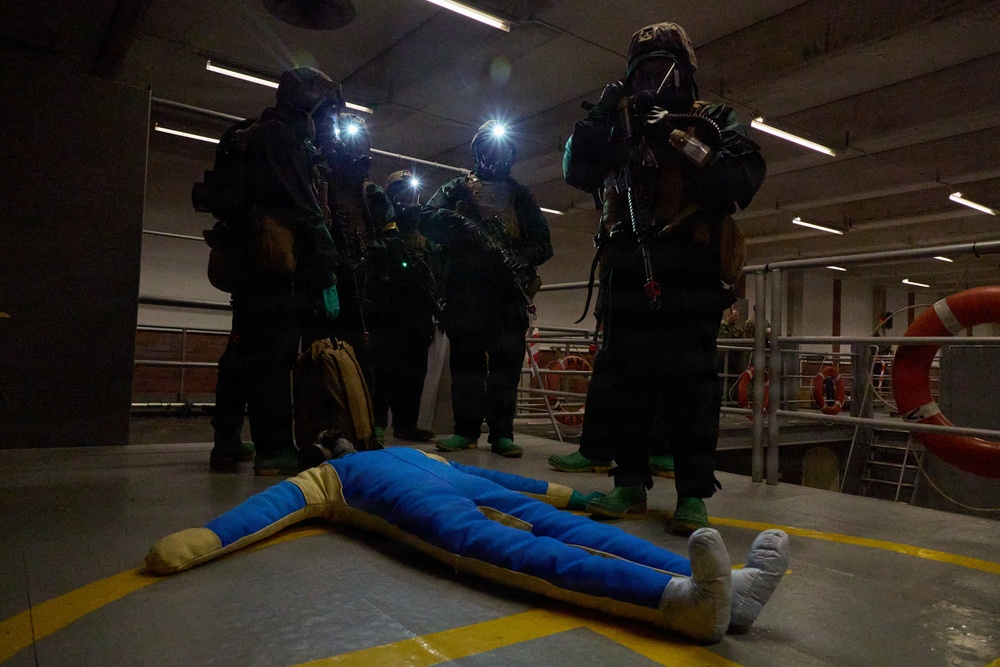 A group of personnel in full CBRN (Chemical, Biological, Radiological, Nuclear) protective gear with gas masks and headlamps are gathered around a training mannequin lying on the floor. The setting appears to be an indoor facility, possibly a ship or industrial building, with life rings visible in the background. This appears to be a military or emergency response training exercise.