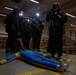 A group of personnel in full CBRN (Chemical, Biological, Radiological, Nuclear) protective gear with gas masks and headlamps are gathered around a training mannequin lying on the floor. The setting appears to be an indoor facility, possibly a ship or industrial building, with life rings visible in the background. This appears to be a military or emergency response training exercise.