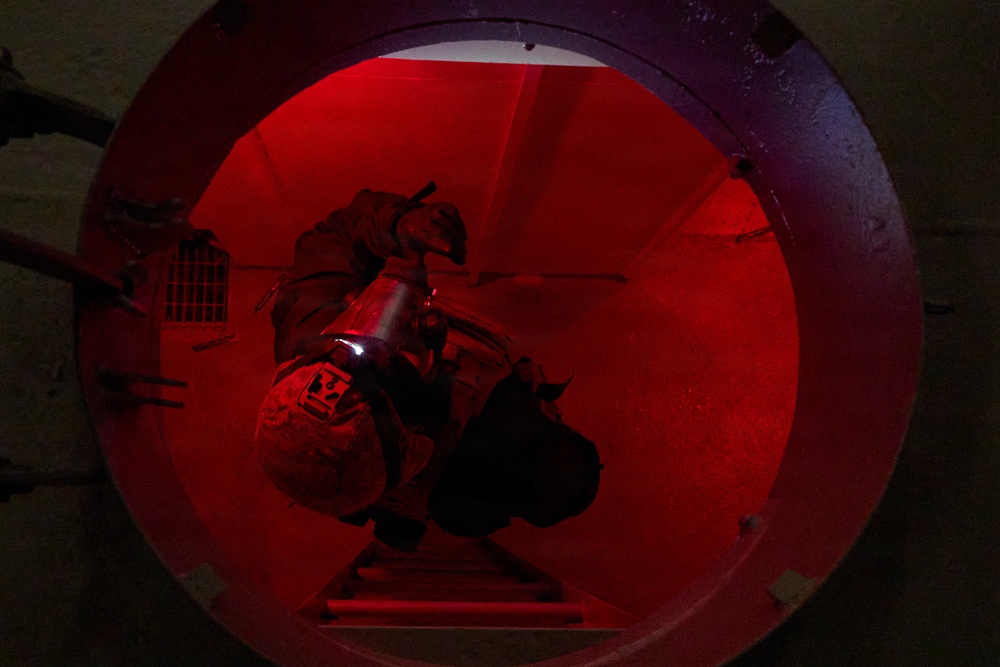A person in military or tactical gear is crawling through a large circular tunnel or pipe illuminated by red light. The individual appears to be carrying equipment including a flashlight or torch, navigating through what appears to be a confined space entry exercise or operation. The circular opening is framed by a thick metal rim, suggesting an industrial or military infrastructure setting.