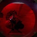 A person in military or tactical gear is crawling through a large circular tunnel or pipe illuminated by red light. The individual appears to be carrying equipment including a flashlight or torch, navigating through what appears to be a confined space entry exercise or operation. The circular opening is framed by a thick metal rim, suggesting an industrial or military infrastructure setting.