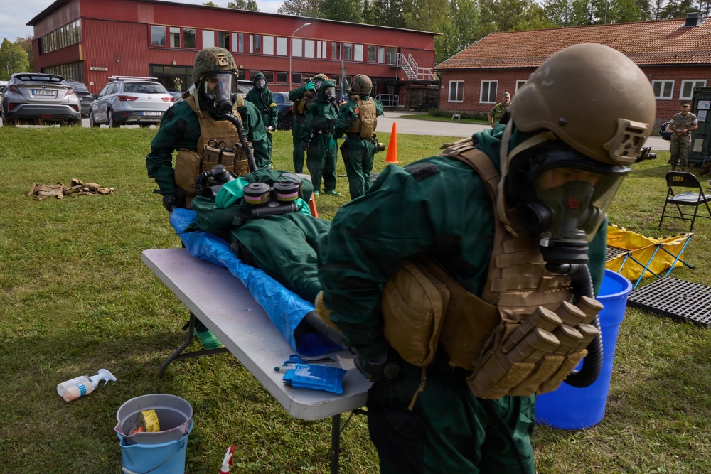 Military personnel in CBRN (Chemical, Biological, Radiological, and Nuclear) protective gear conduct a decontamination training exercise outdoors near a red brick building. Personnel wearing green hazmat suits, gas masks, tactical vests, and helmets are practicing casualty decontamination procedures on a simulated victim lying on a folding table covered with a blue tarp. The setting appears to be a Scandinavian military installation based on the architecture and vehicle license plates.