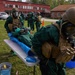 Military personnel in CBRN (Chemical, Biological, Radiological, and Nuclear) protective gear conduct a decontamination training exercise outdoors near a red brick building. Personnel wearing green hazmat suits, gas masks, tactical vests, and helmets are practicing casualty decontamination procedures on a simulated victim lying on a folding table covered with a blue tarp. The setting appears to be a Scandinavian military installation based on the architecture and vehicle license plates.