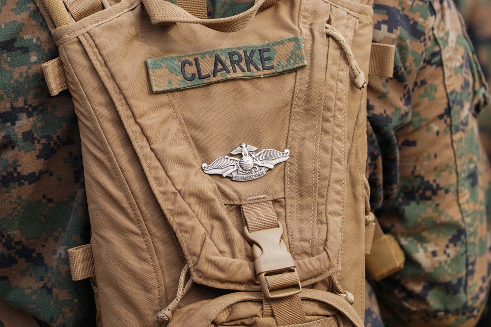 A close-up photograph of a tan/coyote brown military backpack or hydration carrier belonging to someone named 'CLARKE,' as indicated by a camouflage name tape patch. A silver military badge, appearing to be a U.S. Marine Corps insignia (likely a Naval or Marine qualification badge), is affixed to the pack. The background shows individuals wearing USMC MARPAT (Marine Pattern) digital camouflage uniforms.
