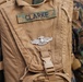 A close-up photograph of a tan/coyote brown military backpack or hydration carrier belonging to someone named 'CLARKE,' as indicated by a camouflage name tape patch. A silver military badge, appearing to be a U.S. Marine Corps insignia (likely a Naval or Marine qualification badge), is affixed to the pack. The background shows individuals wearing USMC MARPAT (Marine Pattern) digital camouflage uniforms.