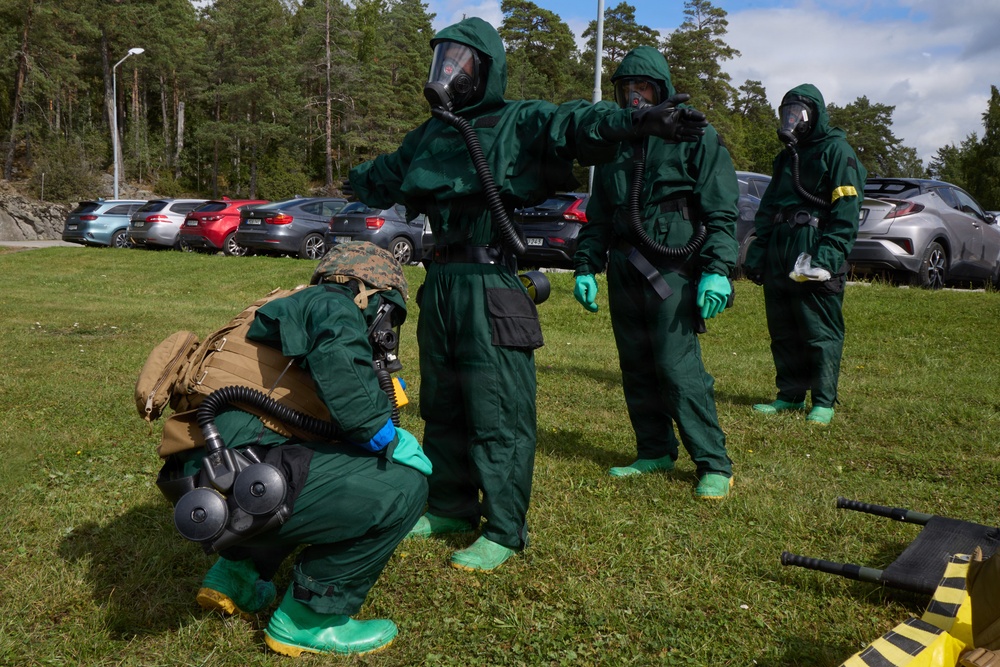 The image shows multiple personnel wearing full CBRN (Chemical, Biological, Radiological, Nuclear) protective suits conducting what appears to be a training exercise outdoors. The individuals are dressed in green hazmat suits with gas masks, green rubber boots, and gloves. The setting appears to be a grassy area near a parking lot in a forested region.