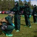 The image shows multiple personnel wearing full CBRN (Chemical, Biological, Radiological, Nuclear) protective suits conducting what appears to be a training exercise outdoors. The individuals are dressed in green hazmat suits with gas masks, green rubber boots, and gloves. The setting appears to be a grassy area near a parking lot in a forested region.