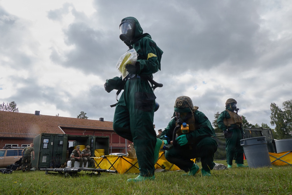 Military personnel are conducting a CBRN (Chemical, Biological, Radiological, and Nuclear) training exercise in an outdoor field setting. Multiple individuals are wearing full green protective suits, gas masks, and green rubber gloves, with yellow decontamination equipment visible on the ground. The setting appears to be a military training facility with red-roofed buildings and shipping containers in the background.