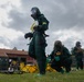 Military personnel are conducting a CBRN (Chemical, Biological, Radiological, and Nuclear) training exercise in an outdoor field setting. Multiple individuals are wearing full green protective suits, gas masks, and green rubber gloves, with yellow decontamination equipment visible on the ground. The setting appears to be a military training facility with red-roofed buildings and shipping containers in the background.